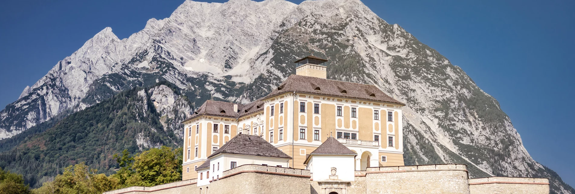 Das helle Schloss Trautenfels erstrahlt gut geschützt von seinen Mauern vor dem anmutigen Grimming, welcher in den wolkenlosen Himmel ragt. | © UMJ